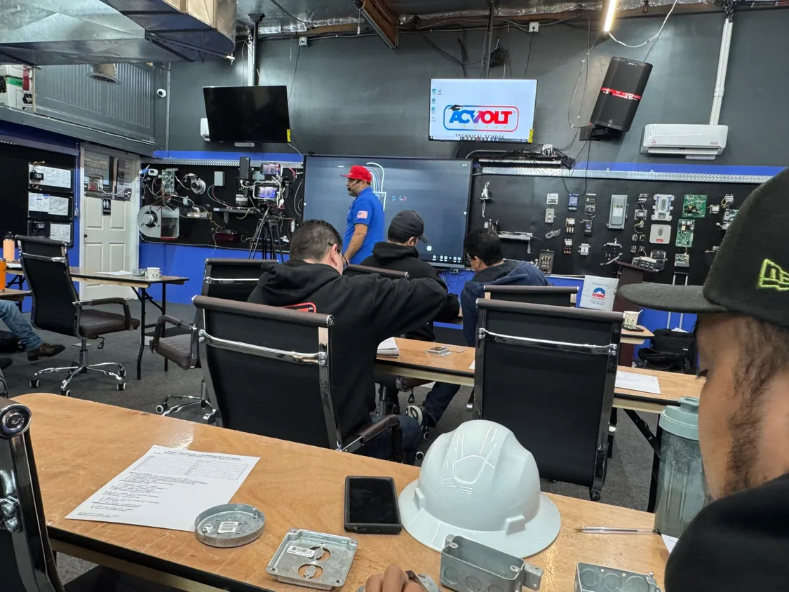 Alumnos sentados en el aula frente al pizarrón digital, con la pared de componentes eléctricos al fondo